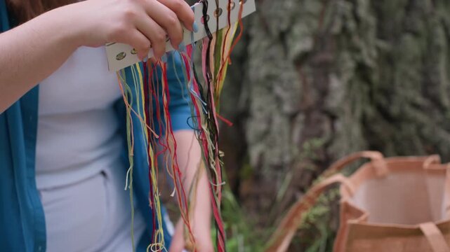 close up of lady holding colorful embroidery threads organized on card while sitting outdoors near tree, preparing for creative needlework session in calm natural setting surrounded by sunlight