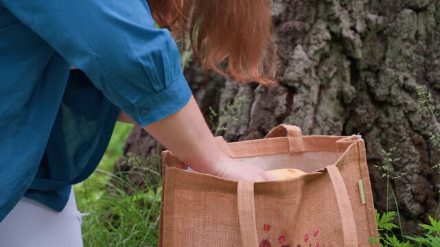 Close up of woman with golden hair returning banana and bottle water into brown fabric bag beside tree trunk, surrounded by green grass and sunlight