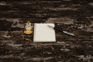 Top view of an open gold pocket watch, a lined notebook, and a metal fountain pen on a weathered, rustic wooden table. Symbolizes writing, planning, time, and history.