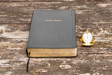 Holy Bible with a black cover and a vintage gold pocket watch on a weathered, rustic wooden table. Symbolizes faith, religion, time, and history.