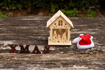 Christmas scene: Miniature wooden house, XMAS letters, and Santa hat on a weathered, rustic wooden table. Outdoor setting in soft daylight. Symbolizes holidays, home, and winter decor.