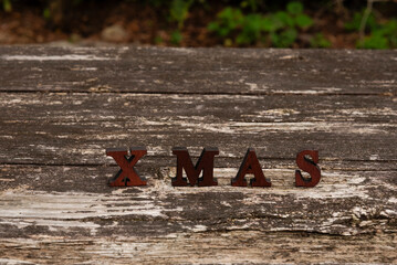 Rustic wooden letters spelling XMAS, placed on a textured, weathered wooden table. Outdoor scene in soft daylight. Symbolizes Christmas, holidays, winter, and handmade decor.