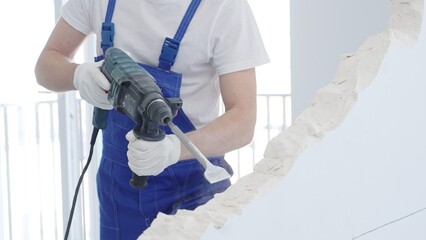 Male construction worker demolishing white wall with rotary hammer drill, wearing blue overalls and...