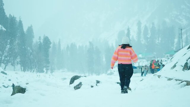 4K Rear view of young woman walks in snow covered mountains during snowfall at Chandanwari, Jammu and Kashmir, India. Female tourist wearing warm clothes exploring snowy winter landscape and enjoying 