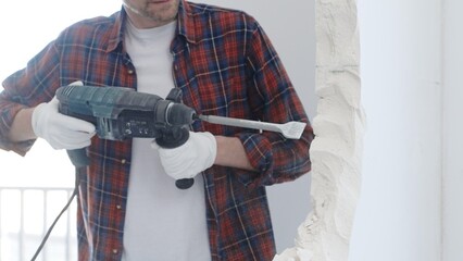 Male construction worker wearing red checkered shirt and protective white gloves is demolishing...