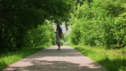 Lady in casual outfit walks backward playfully before turning around to continue strolling along green leafy alley, sunlight filtering through trees, creating peaceful summer - Powered by Adobe