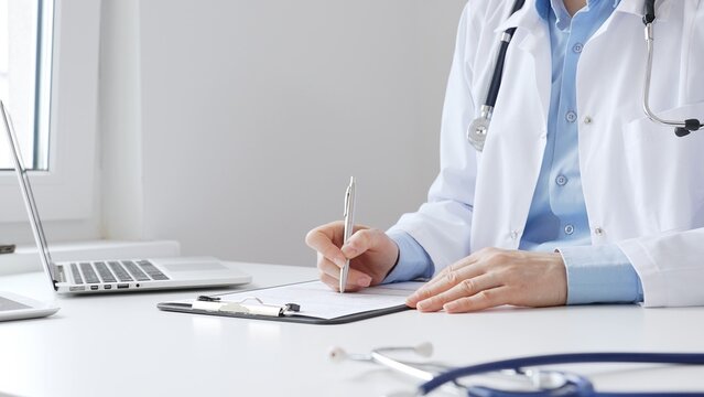 Female doctor completing healthcare paperwork, clipboard positioned near laptop and stethoscope in well lit clinical workspace by window. Medicine and health care concept