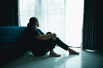 A lonely woman sits on the floor by window looking outside in dark room. Concept of depression,...
