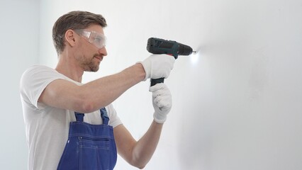 Middle aged man wearing blue overall and protective glasses, is drilling hole in white wall,...