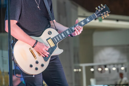 A black-clad musician on a dimly lit indoor stage, playing a white electric guitar Performing an action with the right hand while gripping the neck with the left Potentially singing into an unseen