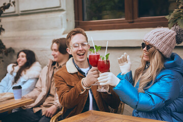 Friends toasting with winter cocktails outdoors in cozy city cafe. Concept of coffee shop advertising, urban lifestyle, and emotional connection during cold season.