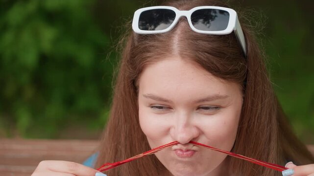 Close up of young woman sitting outdoors holding red embroidery thread across face making funny playful expression , surrounded by lush greenery and soft natural daylight in relaxed atmosphere