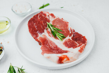 Uncooked pork steak on white plate with rosemary and pepper