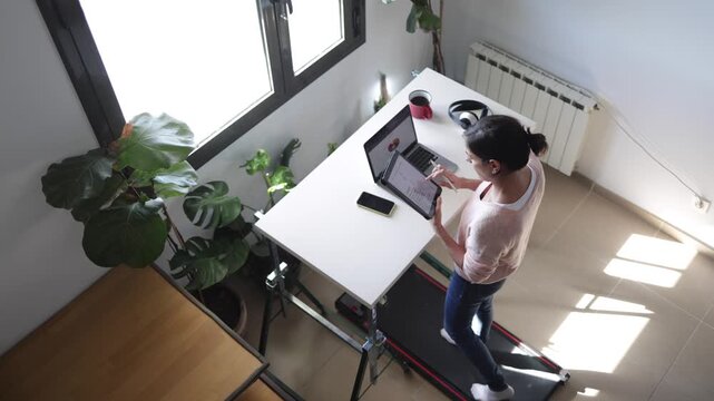 Woman working on a treadmill desk at home, using a laptop and digital tablet, embracing a healthy and active lifestyle while managing remote work and personal well-being