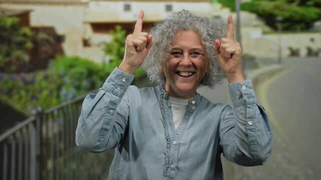 Woman with grey hair smiling and pointing upwards outdoors on a city street with a cheerful expression in an urban environment wearing a blue shirt during daytime.
