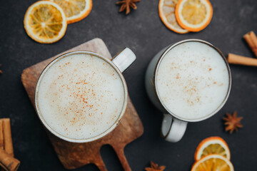 Two cups of spiced latte with cinnamon and orange slices on dark table