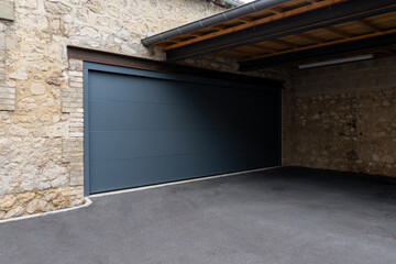 Porte de garage grise anthracite installée par un professionnel, encadrée par un mur en pierre beige et un sol en macadam, entre modernité et authenticité.