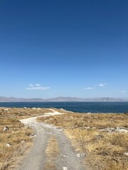 Narrow dirt path through dry grass leading to a blue lake under clear sky