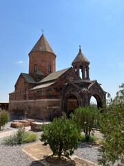 Ancient stone church with conical dome and ornate bell tower surrounded by greenery under clear blue sky