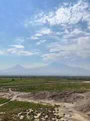 Panoramic view of a vast green plain with rugged terrain and distant mountains partially covered by clouds under a bright blue sky