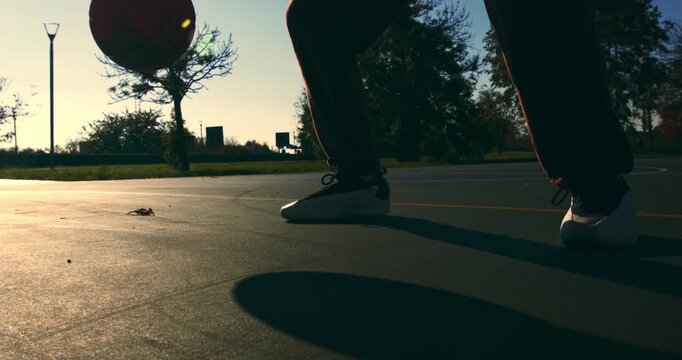 Slow motion close up of young athletic sportsman feet and ball during dribble on asphalt court. Shadows, sunlight, and smooth motion define this street moment.