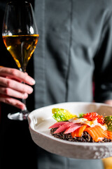 A person holds a gourmet seafood dish with salmon, tuna, quinoa, and roe, and a wine glass. Fine...