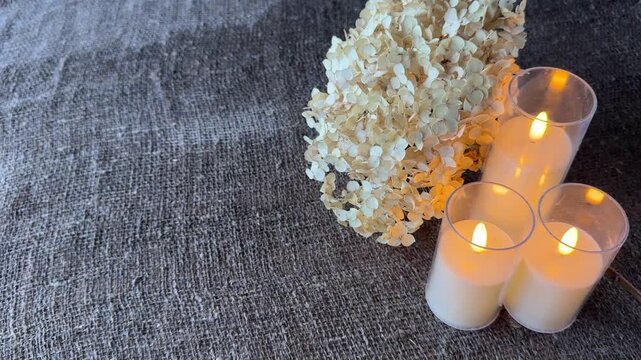 Lush dried hydrangea flower, It lies next to three electric candles with burning wicks, On a surface of rough damp linen burlap