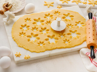 Rolled out dough with cut out Christmas gingerbread snowflake shapes with baking ingredients and with rolling pin on white kitchen table. Top view. Christmas baking concept background.