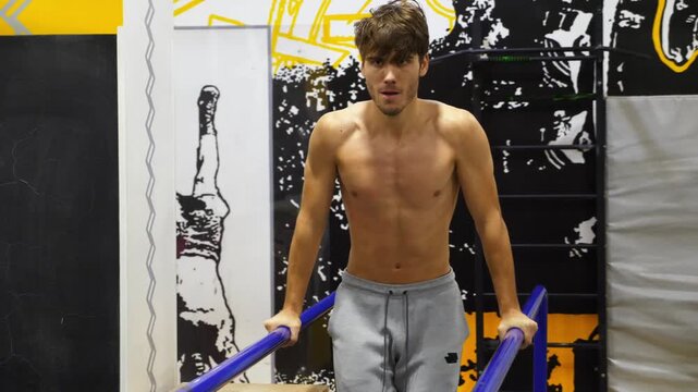 Strong sporty man doing push ups on parallel bars at modern gym. Young sportsman exercising at fitness centre. Male shirtless athlete training hard indoor. Concept of active sportive lifestyle