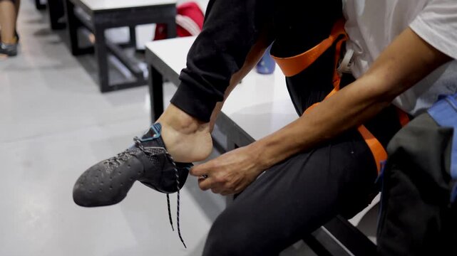 Man in safety harness and black sport pants sits on a bench lacing tight climbing shoes, focused and preparing for an indoor bouldering or rock-climbing session