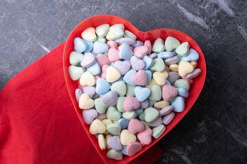Pastel candy conversation hearts in a red heart shaped bowl on marble background with red cloth.  Valentine's Day background
