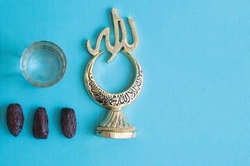 Golden Islamic crescent moon ornament with Arabic calligraphy beside dates and water on a blue background