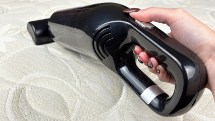 Close-up of a woman's hand vacuuming dust and dirt from a bed mattress with a portable cordless vacuum cleaner. Home cleaning, hygiene, and furniture care concept. Clean house and comfort