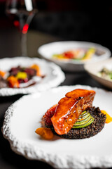 Gourmet close-up of a glazed fish fillet, possibly salmon, served on a bed of dark quinoa with sliced zucchini and a vibrant orange sauce on a textured plate