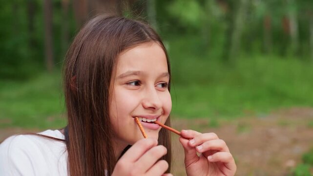 carefree girl playfully putting breadsticks into mouth while smiling during outdoor picnic surrounded by green trees enjoying fun and laughter in warm summer day full of youthful energy