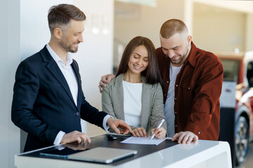 Obraz premium Happy young couple finalizing car purchase at dealership center, signing the contract. Salesman in black suit helping them through the process, showing where to sign, closeup