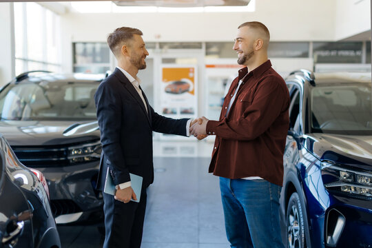 Salesman shaking hands with smiling male customer in a car dealership showroom, greeting with purchase, luxury vehicles displayed around them, happy young man buying new auto - Powered by Adobe
