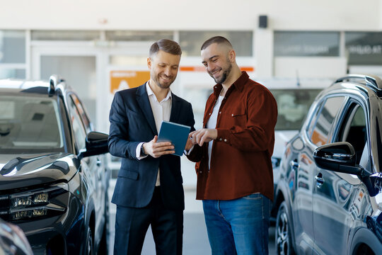 Caucasian millennial salesman showing digital tablet to a male customer, manager in suit and buyer standing between cars in modern dealership showroom, discussing auto characteristics