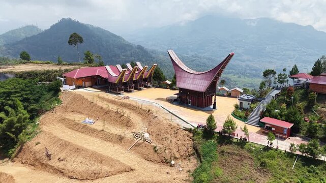 Aerial view of traditional Tongkonan houses with curved roofs on a hillside in Tana Toraja, Indonesia, surrounded by mountains and clouds.	
