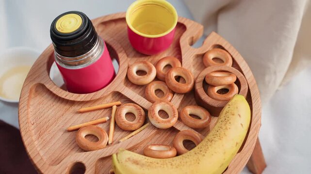 aerial view of small picnic table with wooden tray holding banana, bagels, thermos, and breadsticks while hand reaches to pick one bagel during outdoor picnic setup in warm daylight