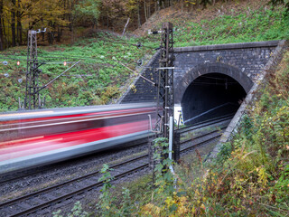Passenger train entering an electrified railway tunnel in autumn forest near Margecany, Slovakia. Motion blur indicates movement; stone portal, overhead lines and double tracks.