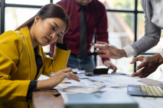 Asian businesswoman stressed while using calculator and reviewing financial reports during a corporate meeting with her colleagues.