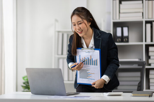 Young office worker having a video call meeting and showing positive results on clipboard with graphs and charts, discussing project details with colleagues online.