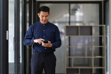Fototapeta premium Businessman using smartphone while standing in modern office corridor, browsing internet, reading news, checking social media, or communicating with colleagues.