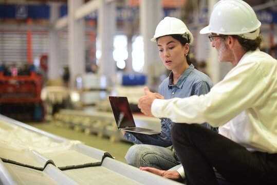 Young Beautiful programmer engineer holding a laptop sitting and discussing with a senior male manager engineer wearing hardhat to solve problems in production line in machinery in industrial plants.