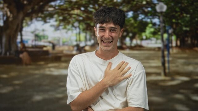 Man with hand on chest laughing in a tree lined street, wearing white t shirt and relaxed posture, visible bare hand; joy sincerity warmth.