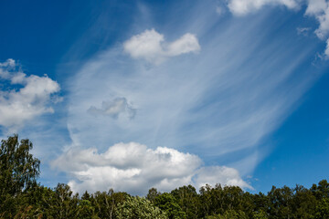 Obraz premium Expansive blue sky showcasing dynamic wispy cirrus and fluffy cumulus clouds over a verdant tree line. A tranquil summer day vista, beckoning adventure