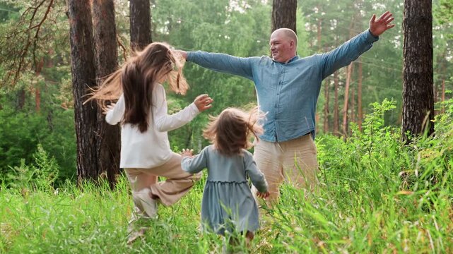 Breadwinner spreading hands wide with joy as daughters run toward him in excitement during cheerful family hangout in green forest, capturing warm emotional reunion moment