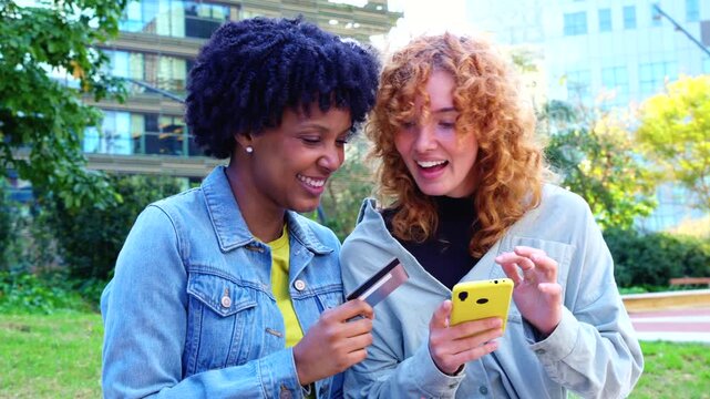 Cheerful African American and Caucasian friends stand in a city park, holding colorful smartphones and a credit card, celebrating cashless convenience, shared shopping success, and digital lifestyle