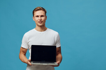 Confident young man with fair hair, wearing casual white t-shirt, holds a closed laptop computer facing camera, standing against bright blue background in studio shot, professional technology concept.